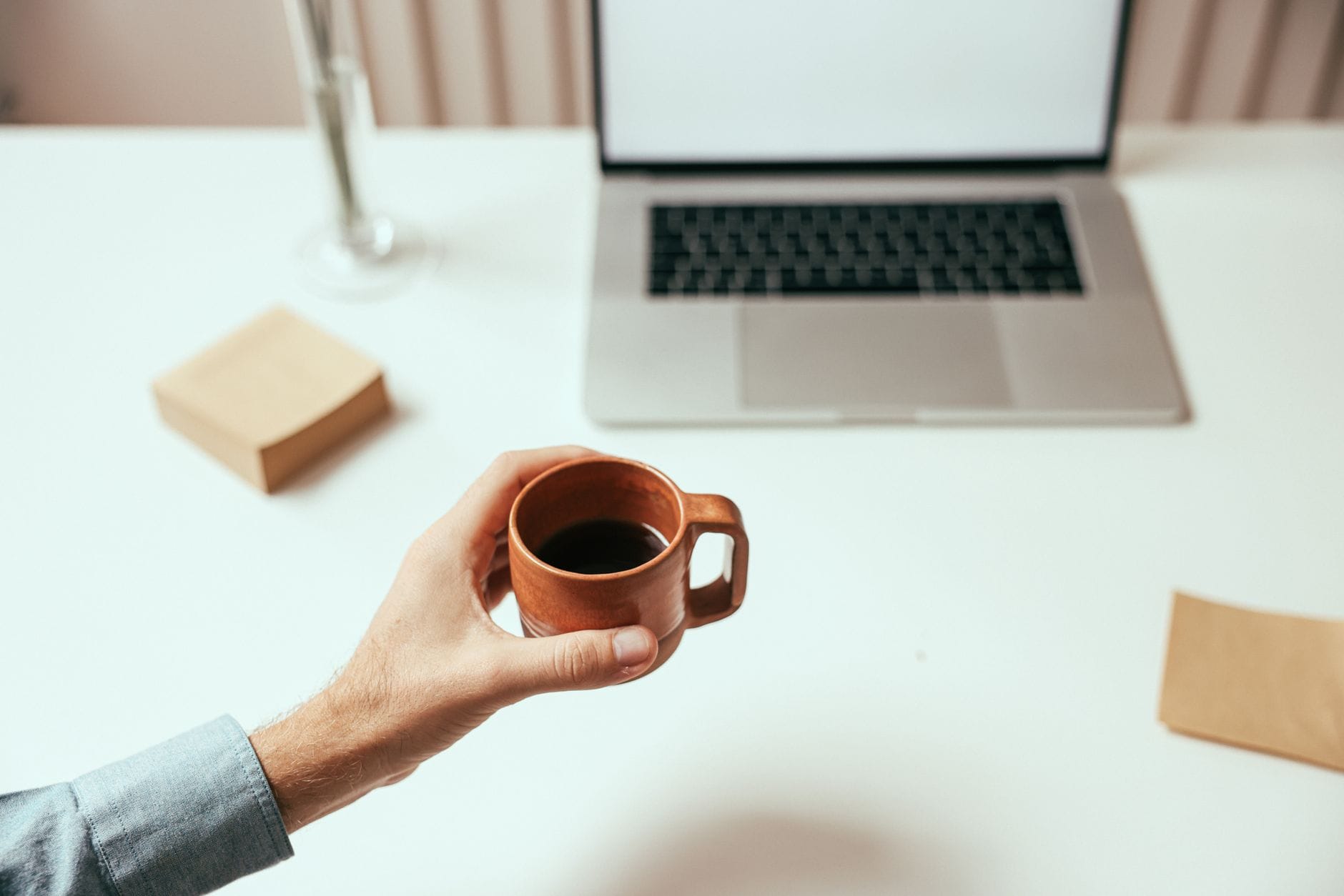 Dłoń z kubkiem kawy obok laptopa na białym, minimalistycznym biurku
